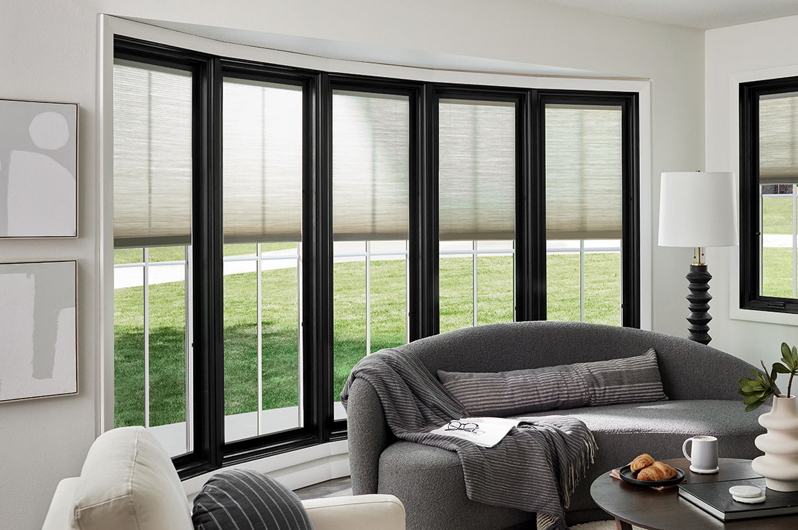 A modern living room featuring a black bow window, a curved gray sofa and a view of the front yard.
