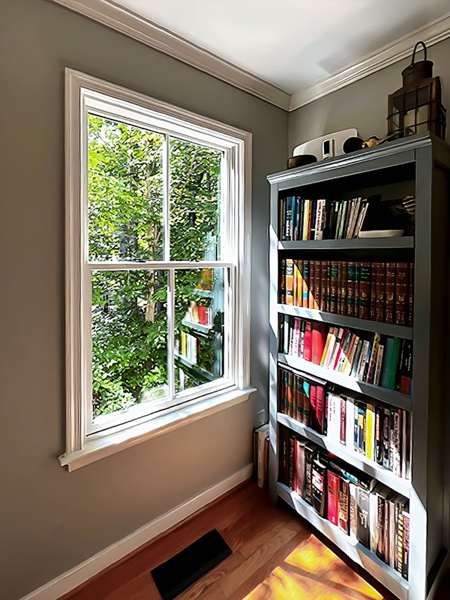 Interior view of a Lifestyle double hung white window with grills next to a bookcase, allowing natural light and outdoor greenery to brighten the reading nook.