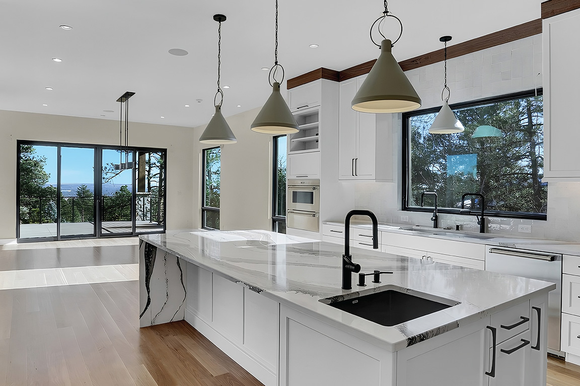 A modern kitchen with white cabinets, a marble island, and large kitchen windows offering a scenic view.