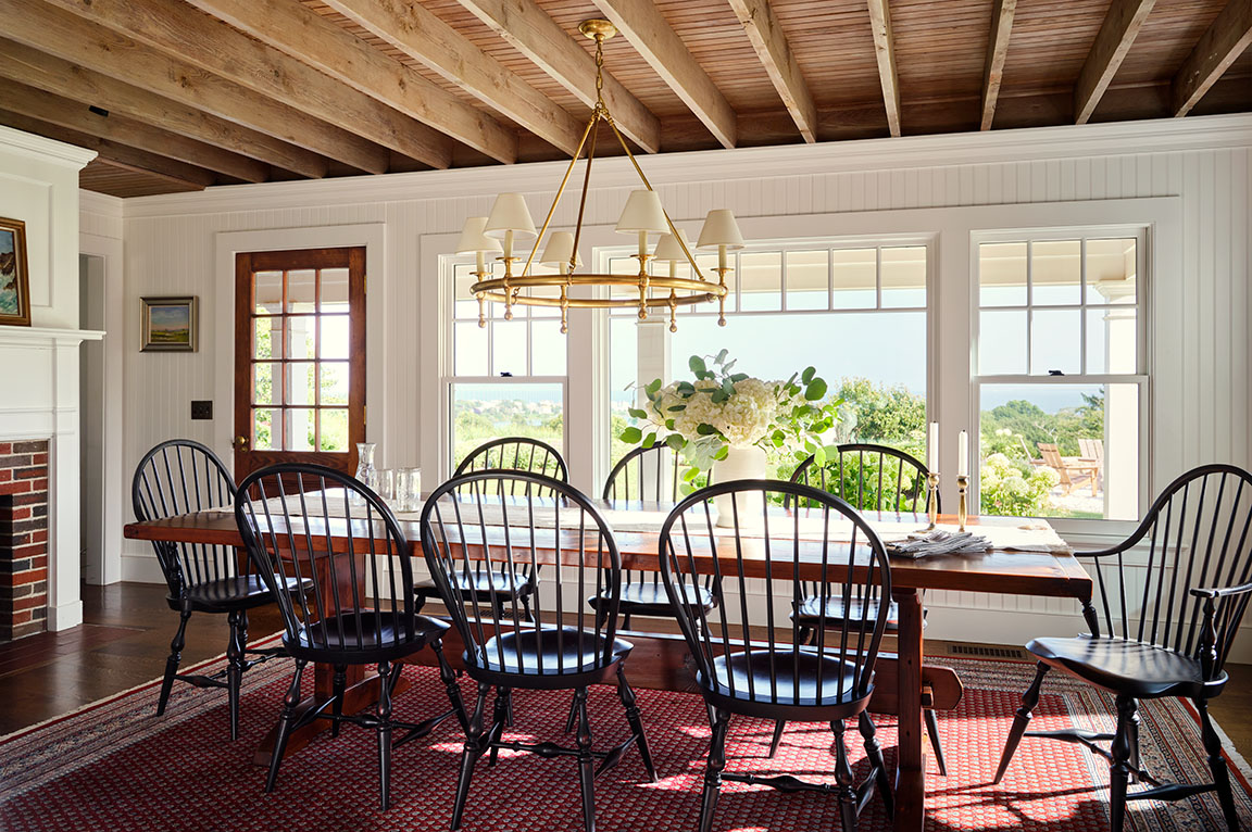 A dining room featuring black chairs around a wooden table, with large windows showcasing a scenic view.