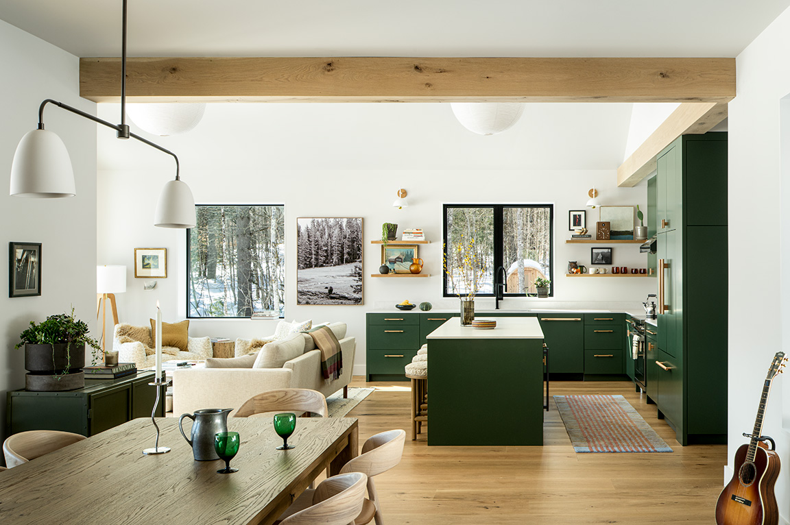 A modern kitchen with green cabinets and a cozy living area, featuring large fiberglass windows overlooking a snowy forest.