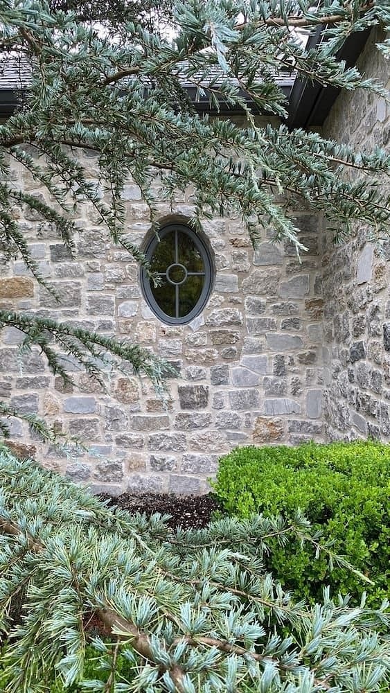 A stone home exterior has an oval window with custom gray grilles. 