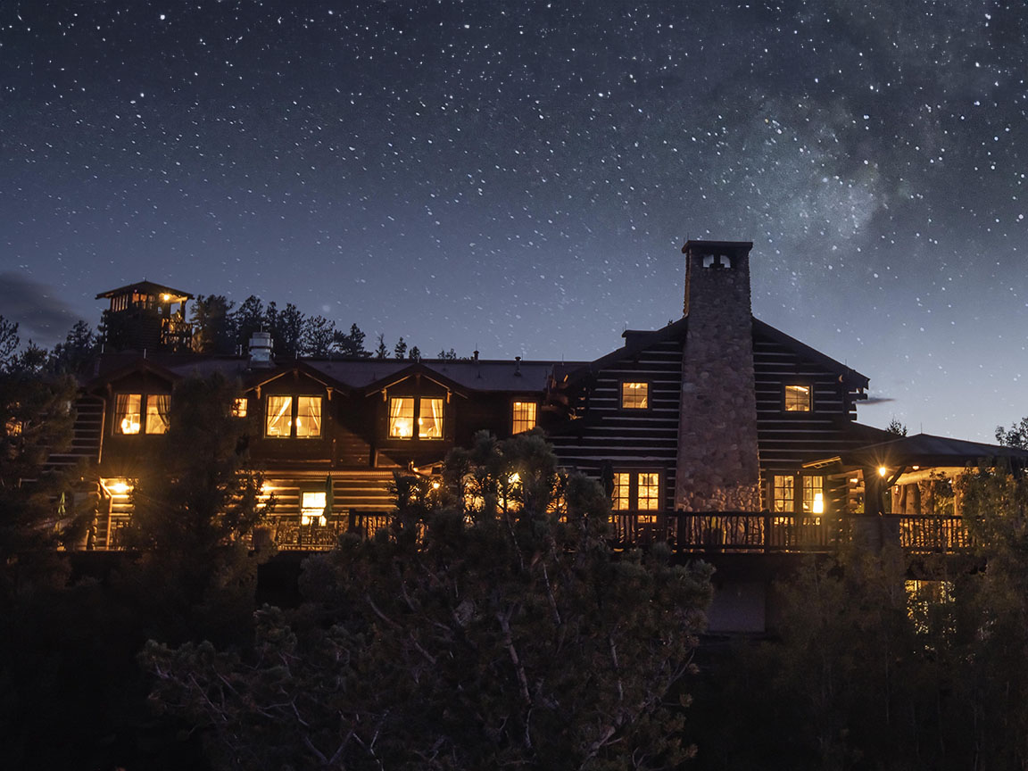 A large log cabin with glowing cabin windows under a starry night sky and surrounded by trees.