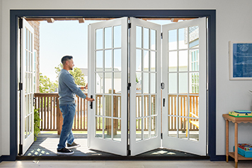 A man wearing blue jeans and a blue shift is opening a four-panel white bifold door.