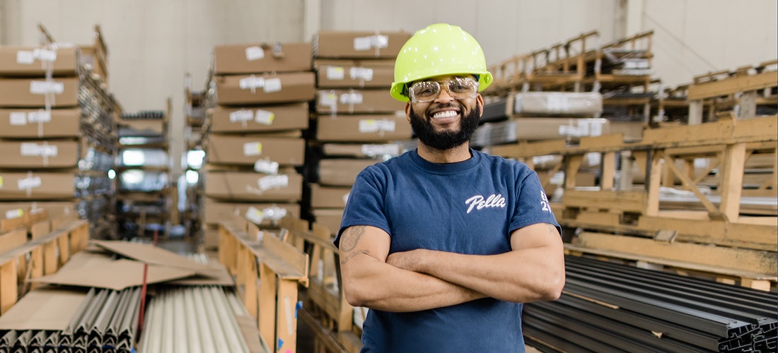 a Pella team member with a yellow hard hat and beard proudly standing in front of many palettes of wood