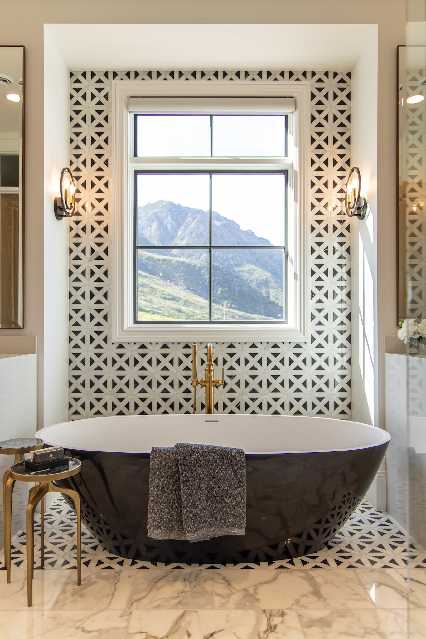 This freestanding bathtub with gold hardware sits on patterned tile with a picture window above it.