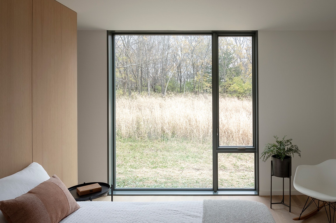 A bedroom with floor-to-ceiling windows overlooking tall grass and trees, letting in natural light.
