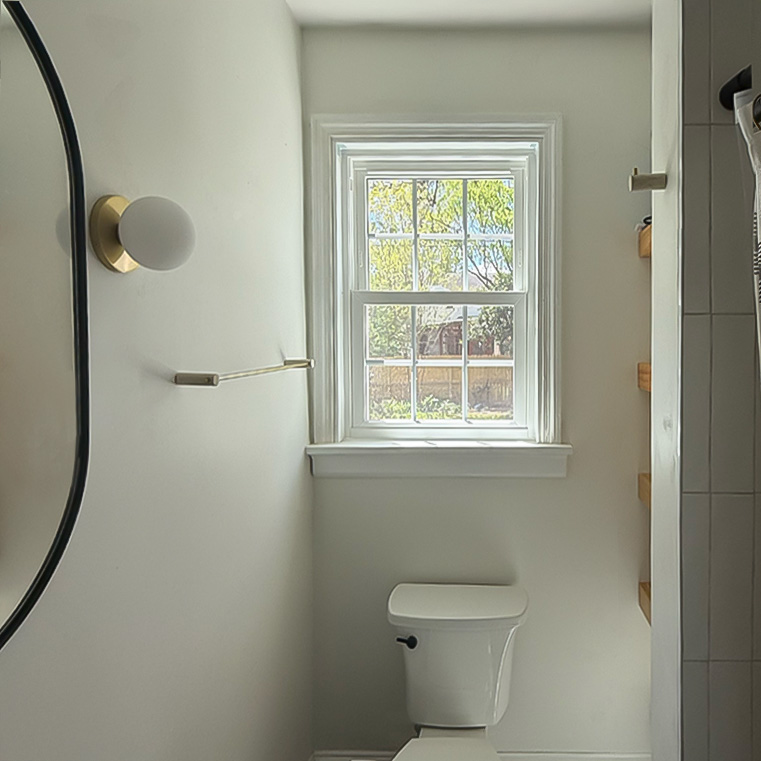 A bright bathroom featuring a window with a view of greenery outside, modern light fixture, towel rack, and a white toilet. The walls are painted light gray, and there is a partial view of shelving on the right side.