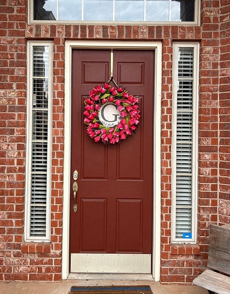a solid front door with a reddish paint and a wreath with a large "G" inside the flowers