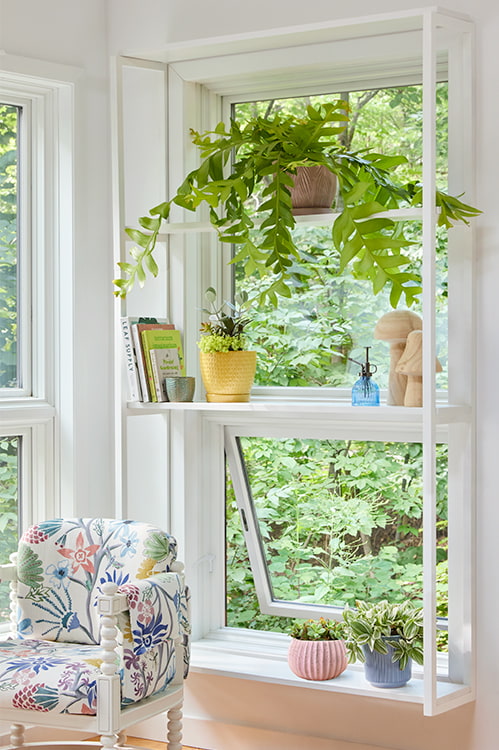 three shelves with several potted plants and other decor items in front of a window that is beside a colorful corner chair.