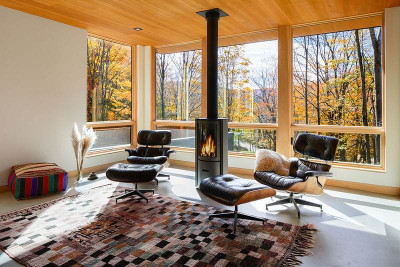 A sunroom with wood windows and a fireplace overlooks an autumn forest outside.