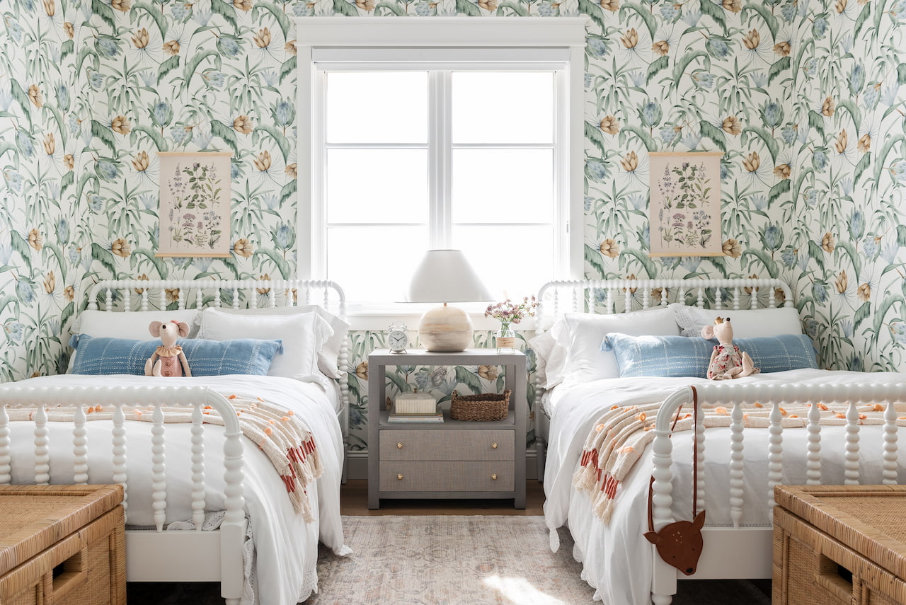This kids' bedroom has patterned wallpaper and two twin-size beds with a white window between them.