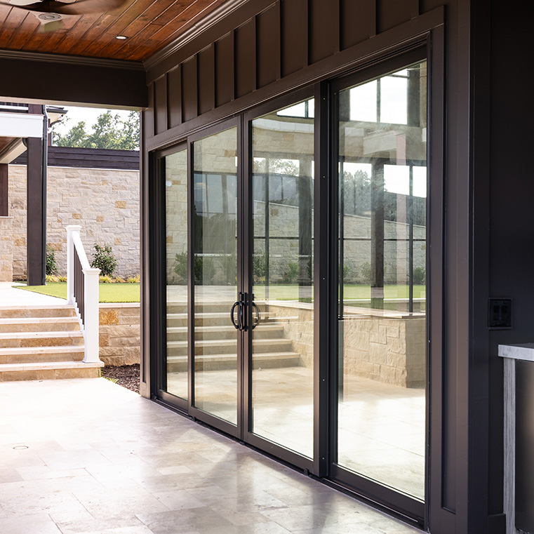 Modern patio with large black-framed sliding glass doors reflecting a stone wall and steps leading to a grassy area. The space features a wooden ceiling with recessed lighting and light-colored tile flooring, showcasing a seamless indoor-outdoor transition and contemporary design.