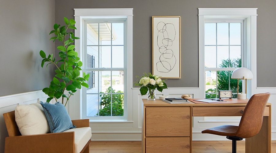 A modern office featuring large windows, a wooden desk, a plant, and decorative art, showcasing natural light.