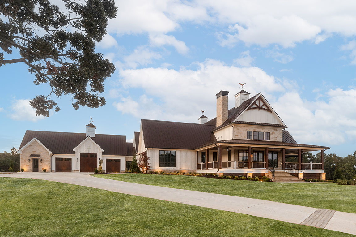 Exterior view of a large modern farmhouse with a metal roof, stone chimney, and wrap-around porch set against a backdrop of green lawn and very blue sky.