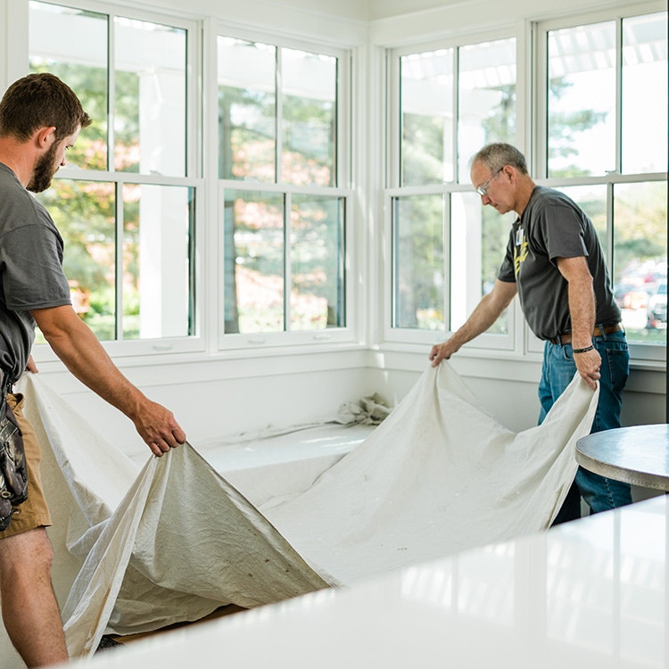 two contractors are preparing to install new windows in a home by laying down protective tarps on the floor.