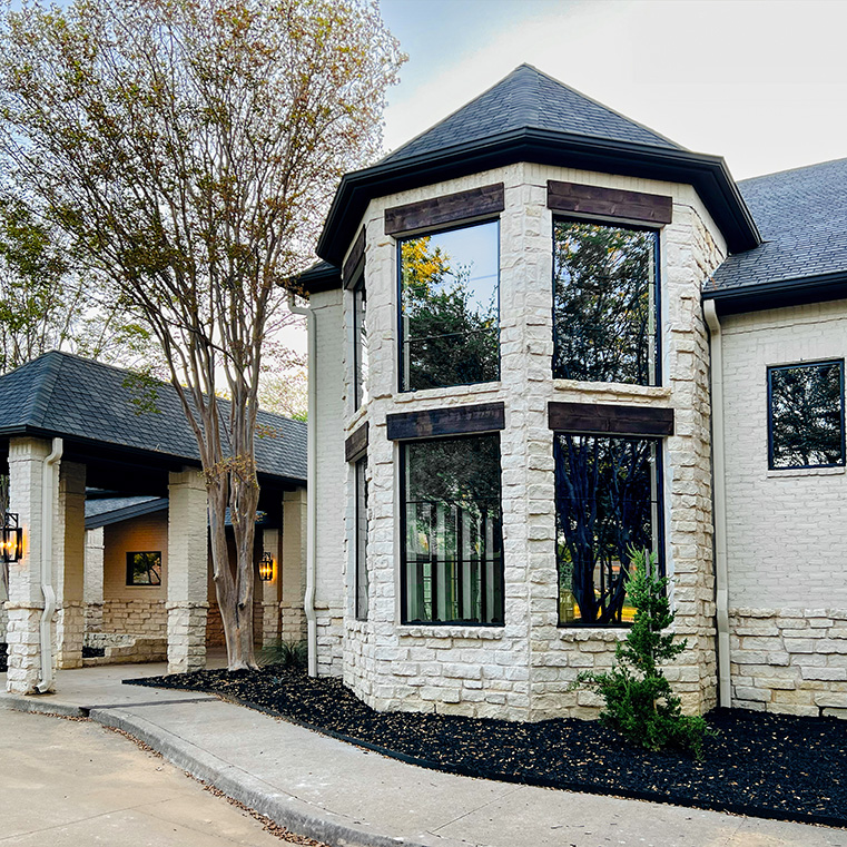 A modern home exterior featuring large black windows and a stone wall, surrounded by landscaping.