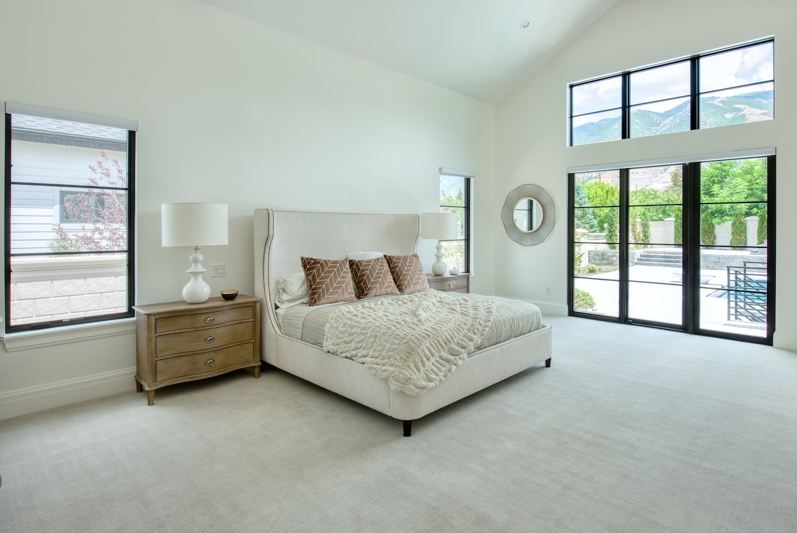 A large bedroom in Utah has a neutral-colored bed positioned between two casement windows and a black sliding glass door on the far wall.