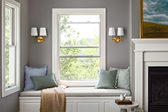 a window bench with various green-colored pillows sits below a white double-hung window that has the bottom sash open.
