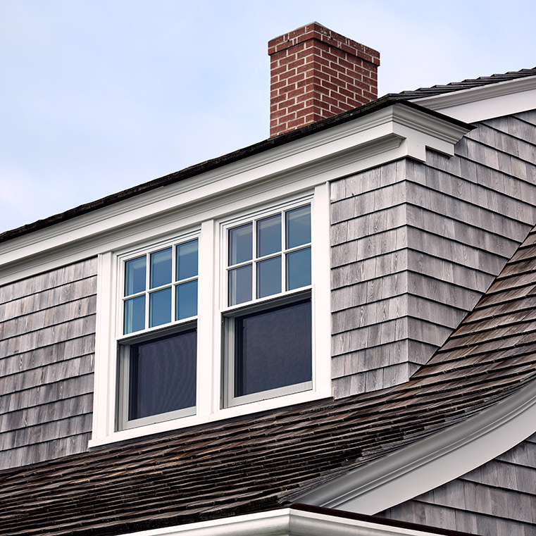 A double-hung window on a shingle roof with a brick chimney against a clear sky.