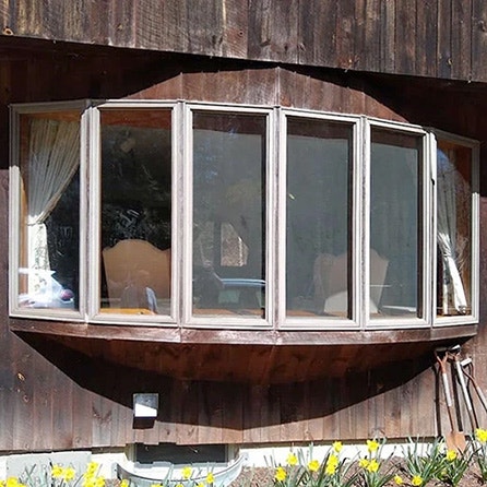 Before white bow window on a wooden house exterior, showcasing large glass panes.