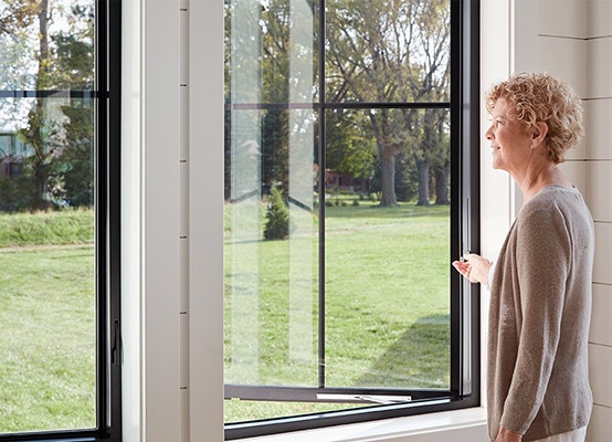 a woman opens her Pella Impervia fiberglass casement window with the innovative easy-slide operator instead of the traditional crank hardware.