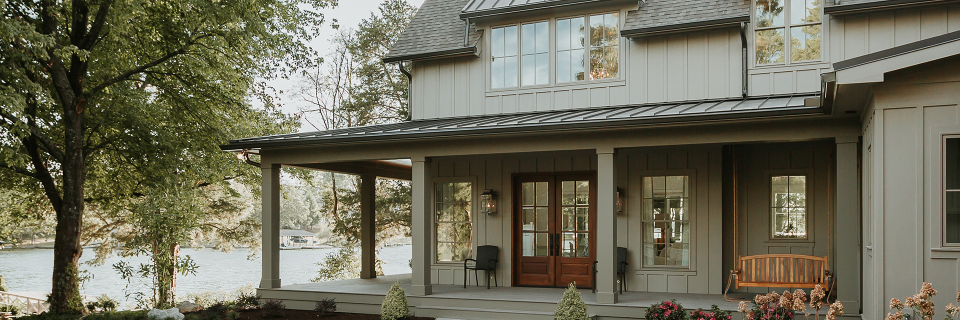 Lakefront home with gray board-and-batten siding, wood front door, large windows, and covered porch with swing.