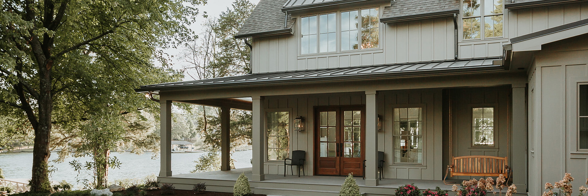 Lakefront home with gray board-and-batten siding, wood front door, large windows, and covered porch with swing.