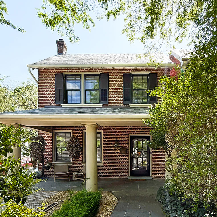 A charming brick house with black shutters, a covered porch, and lush greenery surrounding it
