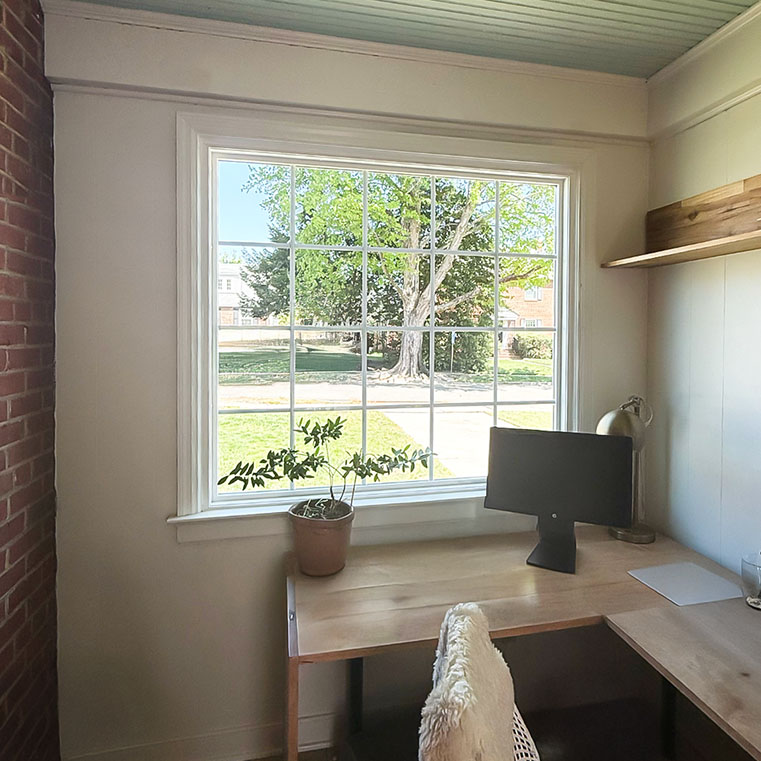 A home office setup featuring a wooden desk with a computer monitor, a potted plant, and a lamp. The desk is positioned in front of a large window that offers a view of a green lawn and trees outside