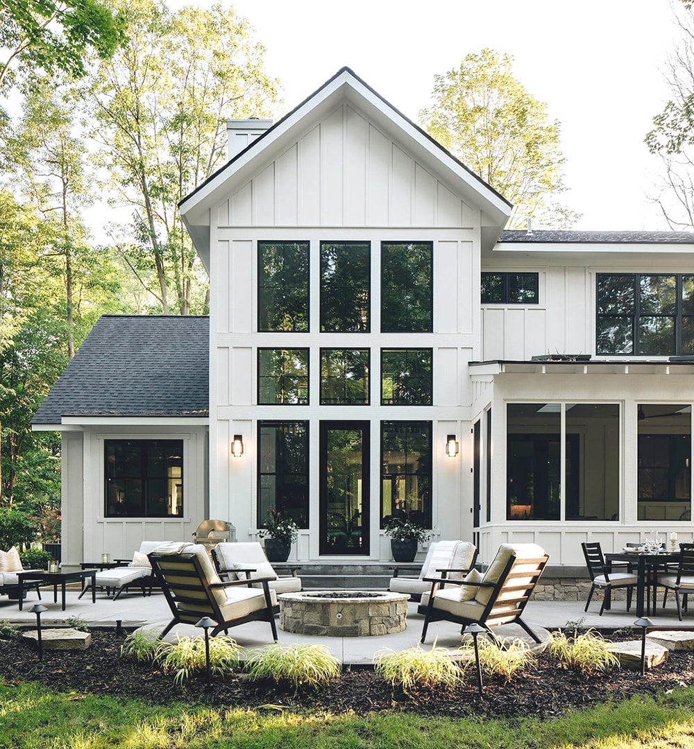 The exterior of a white farmhouse has black picture windows and a black hinged patio door, opening to a fire pit patio.