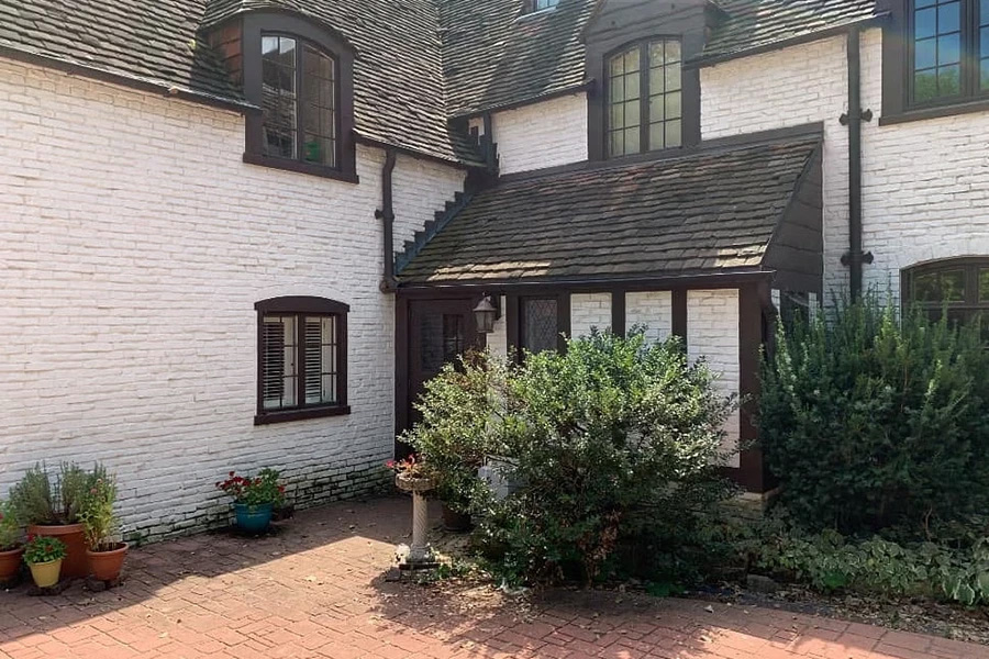 A brick courtyard leads up to a white historic home with large shrubbery in front of the windows.