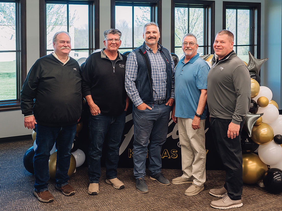 Five people stand in front of large windows with balloons and outdoor scenery visible through the glass.