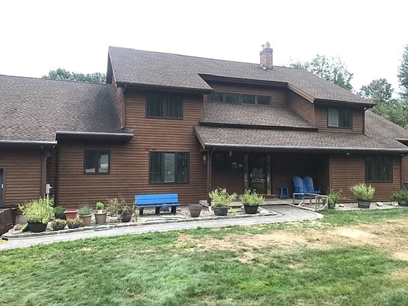 A brown wooden house with front windows before, featuring a blue bench and potted plants in the yard.