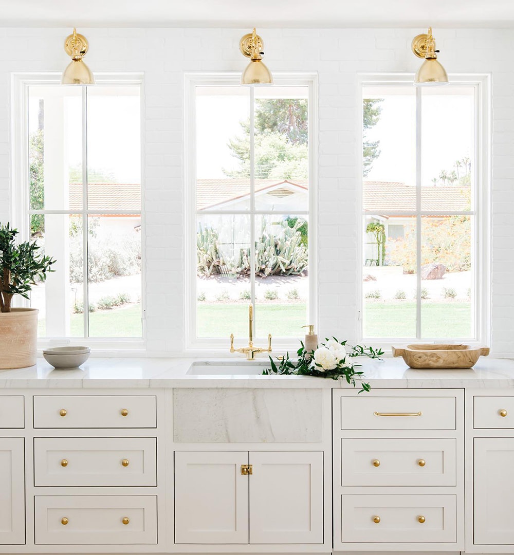 White kitchen with gold hardware features three traditional windows with simple grilles over the sink.