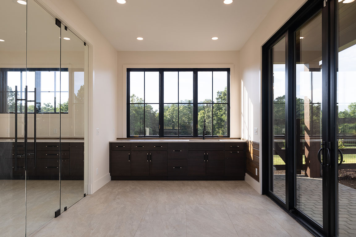 Modern interior space with large windows and glass doors, featuring dark wooden cabinetry beneath a wide window overlooking greenery. The room has light stone tile flooring, recessed ceiling lights, and a minimalist design that emphasizes natural light and openness.
