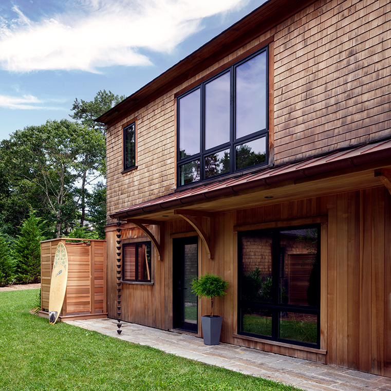 A cape cod house with black windows and wooden siding, featuring a grassy yard and a surfboard.