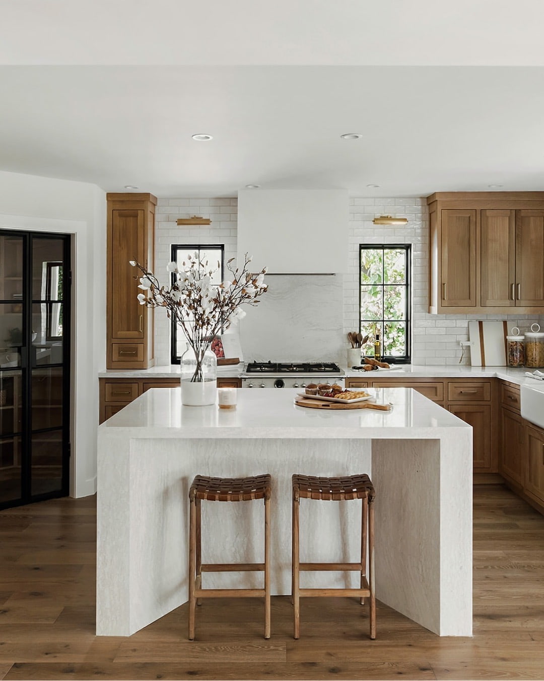 A kitchen with wood cabinets has black windows on both sides of the stove with art lights hung above.