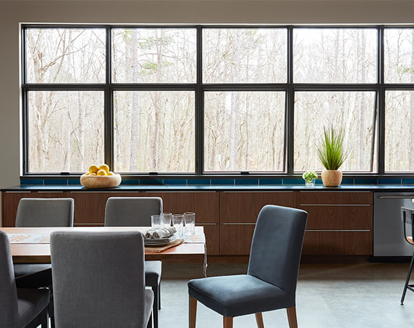 A contemporary dining room table in front of a row of Pella Reserve awning windows