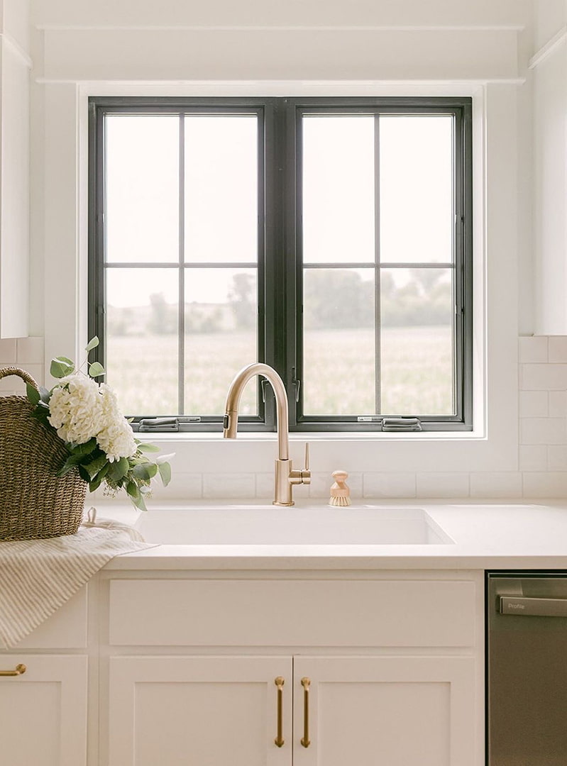 A white kitchen with gold hardware has two black casement windows over the sink.