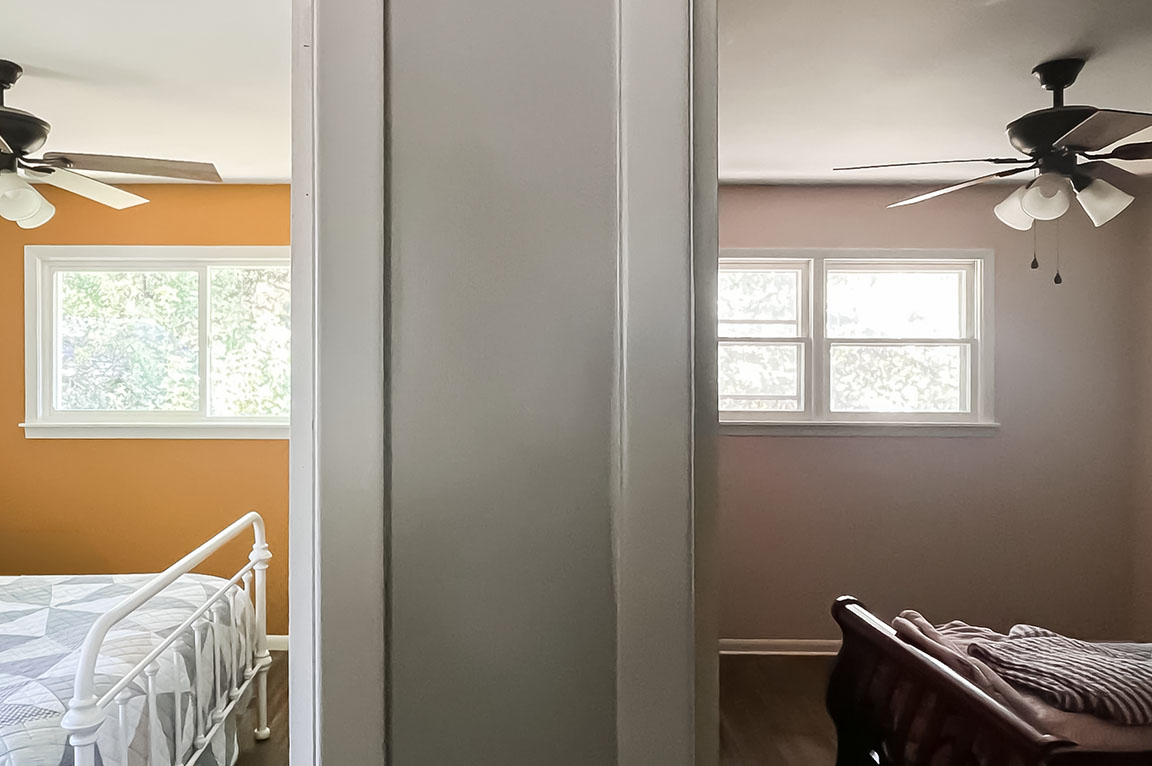 Two adjacent bedrooms separated by a narrow wall — left: orange wall with a white metal bed; right: pink wall with a dark wood sleigh bed, windows and ceiling fans