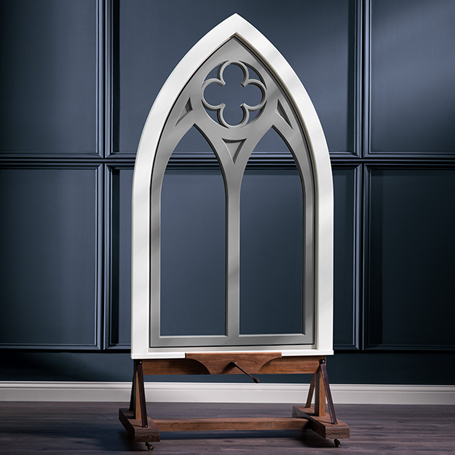 A gothic arched window design in a white frame, displayed on a wooden stand against a dark wall.