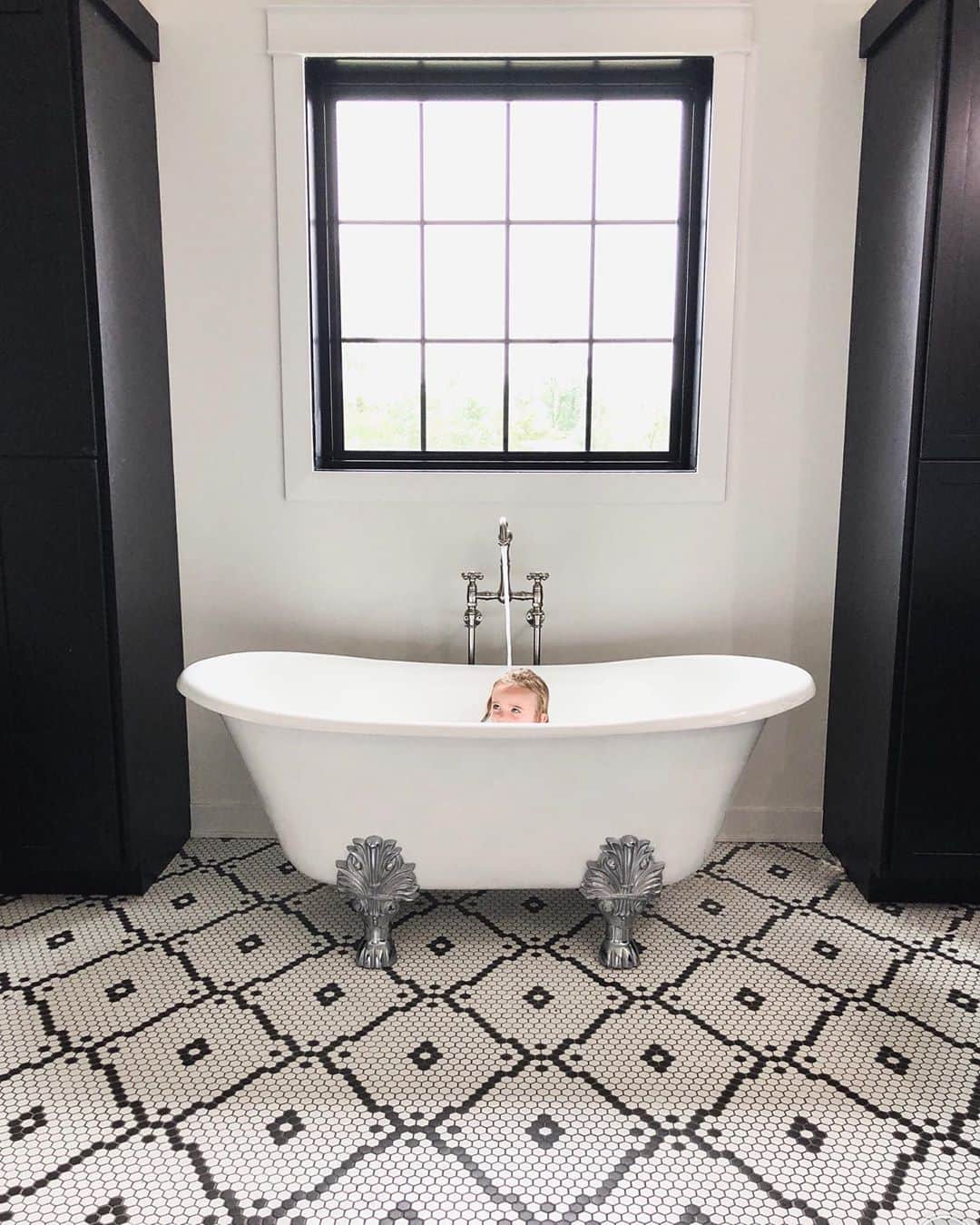 black and white bathroom with face peeking out of the freestanding tub and black picture window directly above