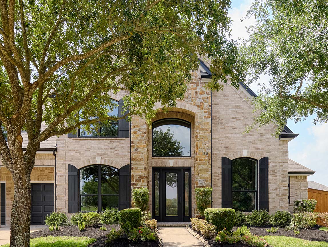 Stone exterior home with large black windows, arched entryway, and landscaped front yard framed by mature trees.