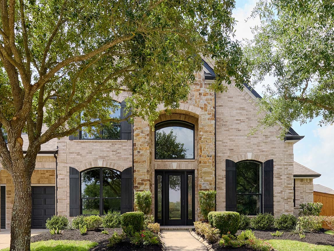 Stone exterior home with large black windows, arched entryway, and landscaped front yard framed by mature trees.