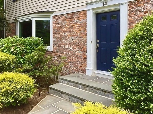 A charming brick house with a blue door and a wood bay window, surrounded by lush greenery.