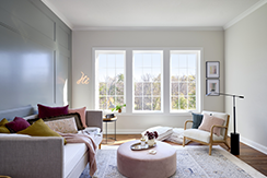 a living room with a dark accent wall on the left and a cream wall on the right has three white casement windows on the far side of the room.
