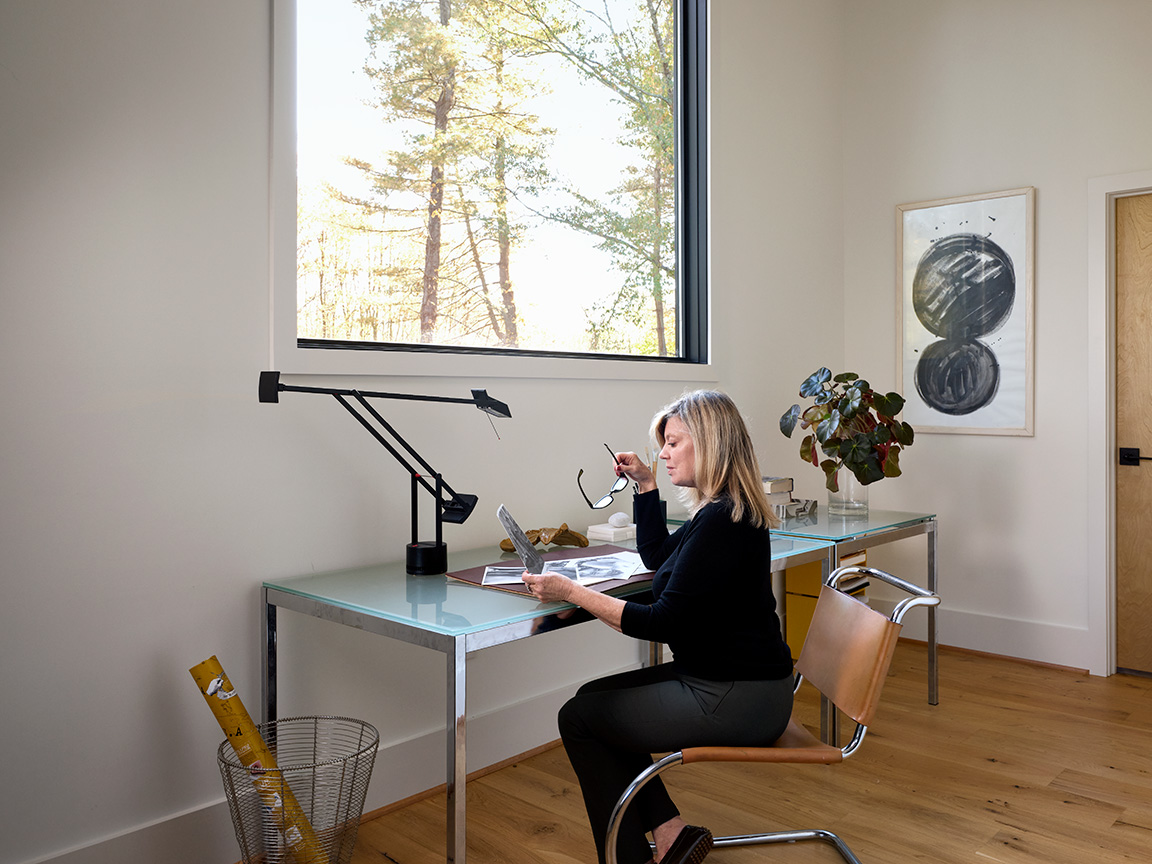 Modern home office featuring a glass desk with a sleek black lamp, wooden chair, and decorative artwork on the wall, with natural light streaming through a large window overlooking trees.