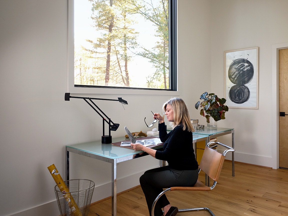 Modern home office featuring a glass desk with a sleek black lamp, wooden chair, and decorative artwork on the wall, with natural light streaming through a large window overlooking trees.