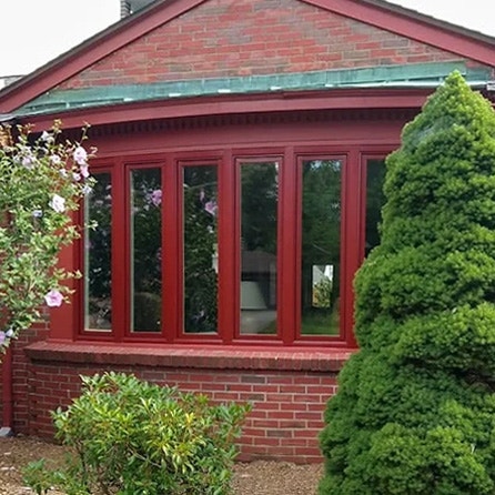 A red bow window replacement on a brick house, surrounded by lush greenery.